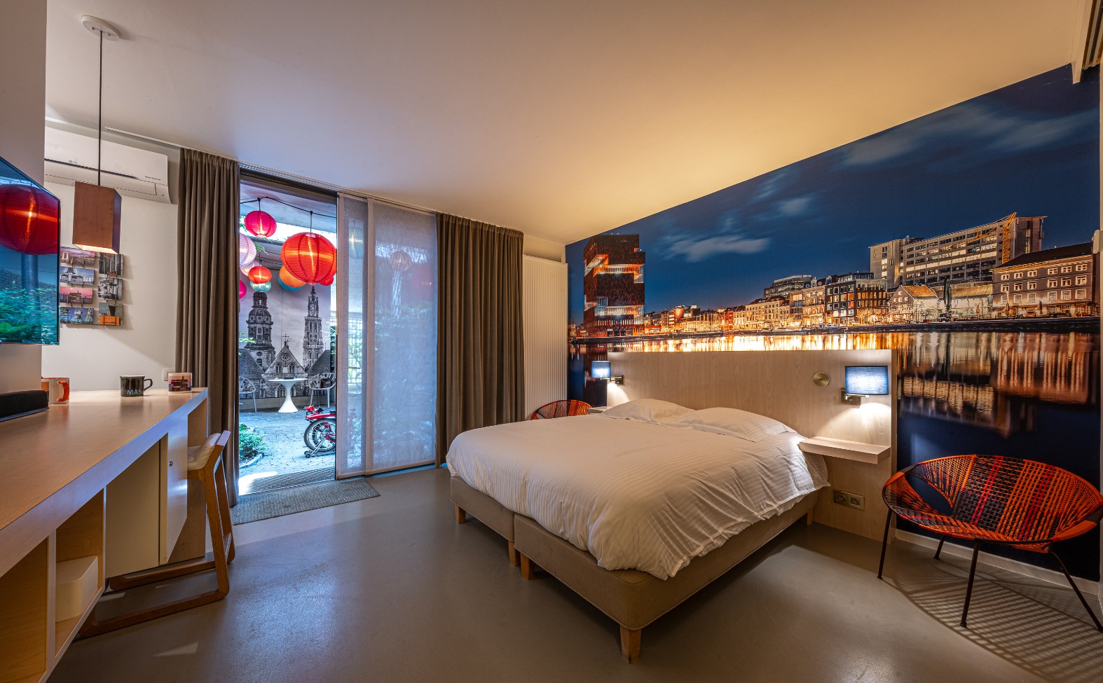 Modern hotel bedroom suite with workspace, balcony access, and illuminated city waterfront views.