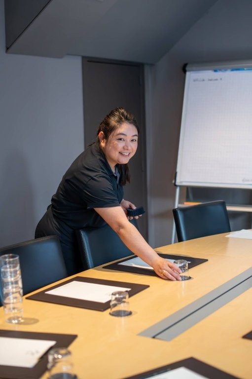 Vrouw in donker uniform aan een moderne vergadertafel met zichtbare waterglazen en notitieblokken.