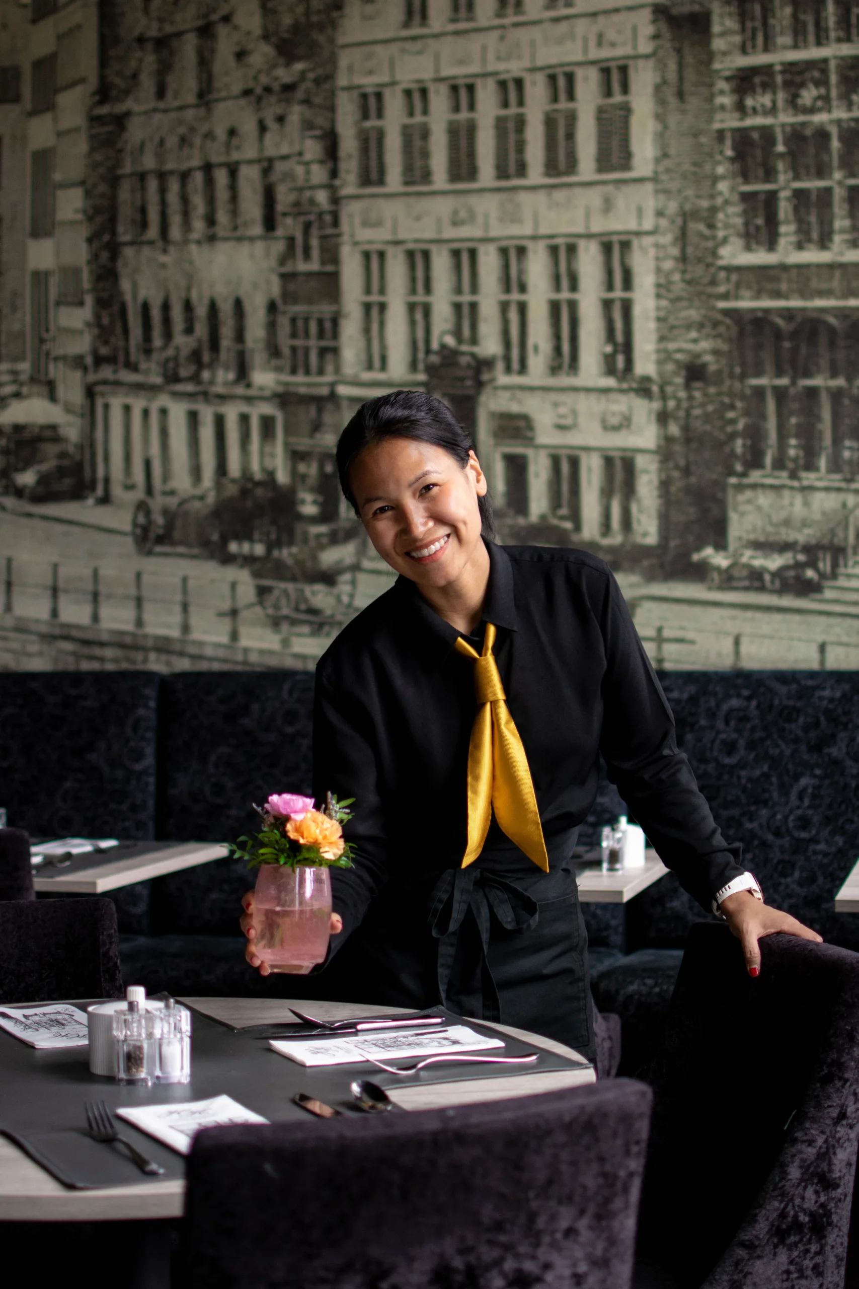 Professioneel geklede vrouw bij de receptiebalie van een elegante lounge met kunstwerken van het stadsgezicht op de achtergrond.