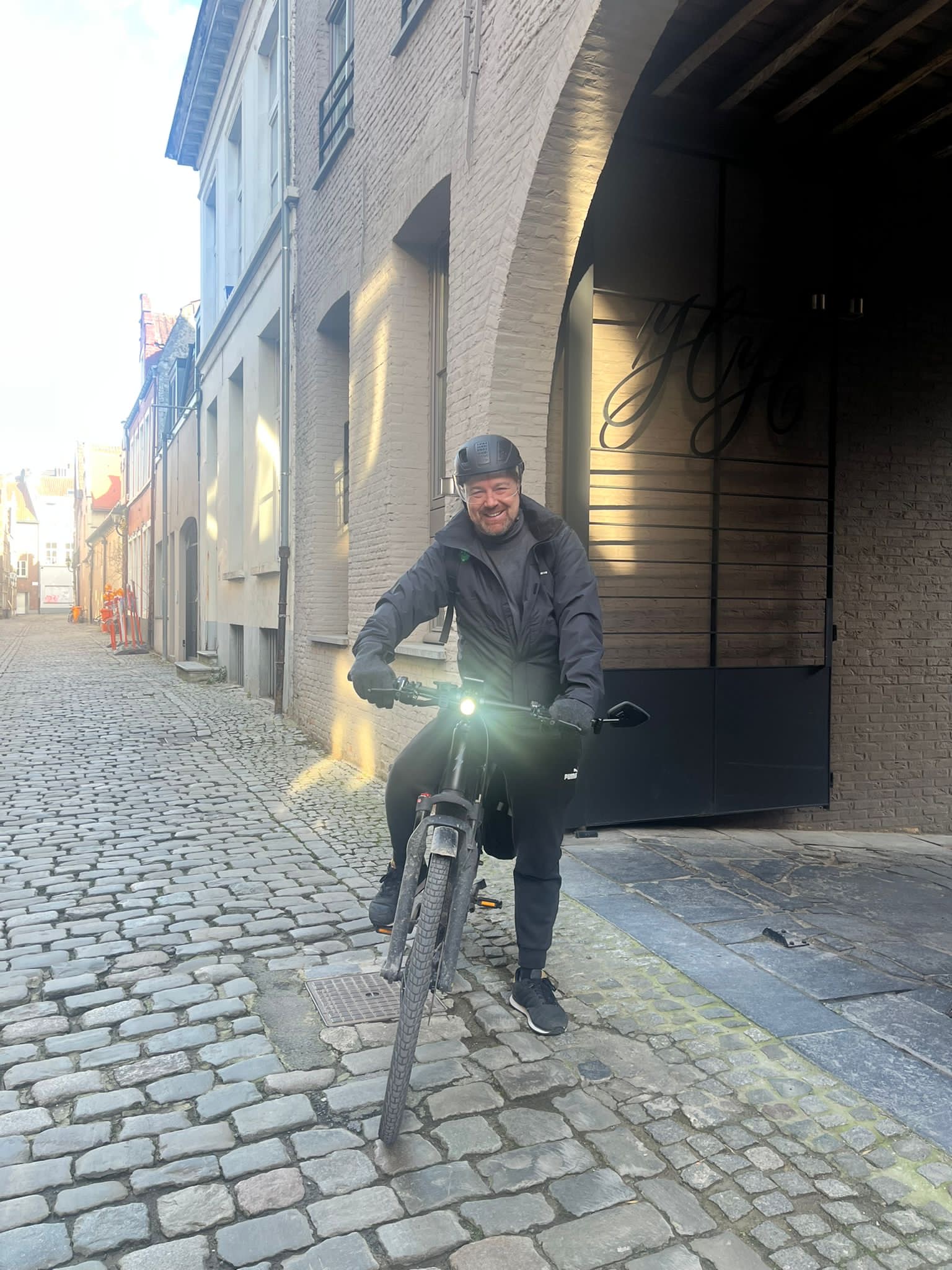 Man op elektrische fiets poseert voor de ingang van een bakstenen gebouw in een Europese straat met kinderkopjes.