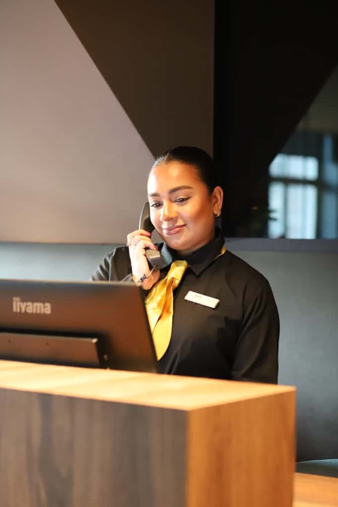 Hotelreceptionist met zwart uniform en gouden stropdas bij de receptie.