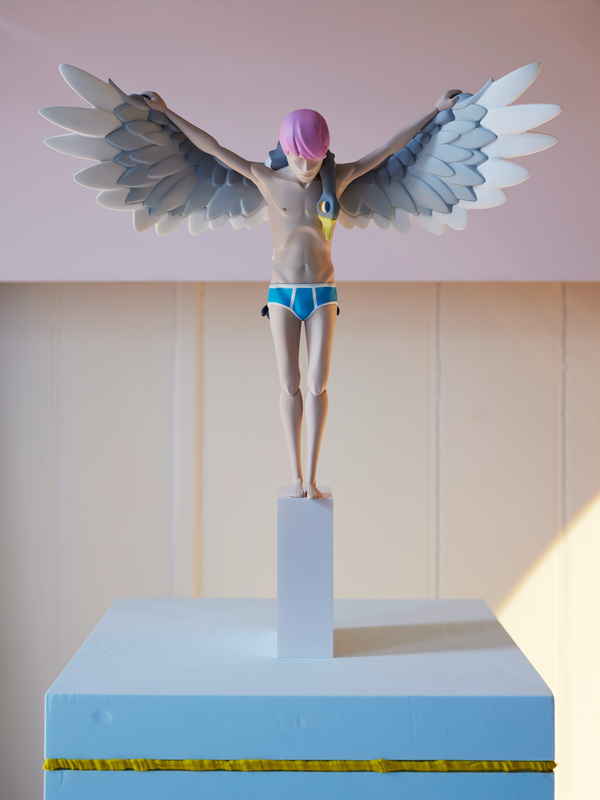 Eine Figur in Menschenform mit rosa Haaren steht auf einem weißen Sockel vor einem hellen Hintergrund. Die Figur hat blaue Badehosen an und ausgebreitete, weiße Flügel. Die Arme sind seitlich ausgestreckt. Die Figur wirkt fragil und nachdenklich.