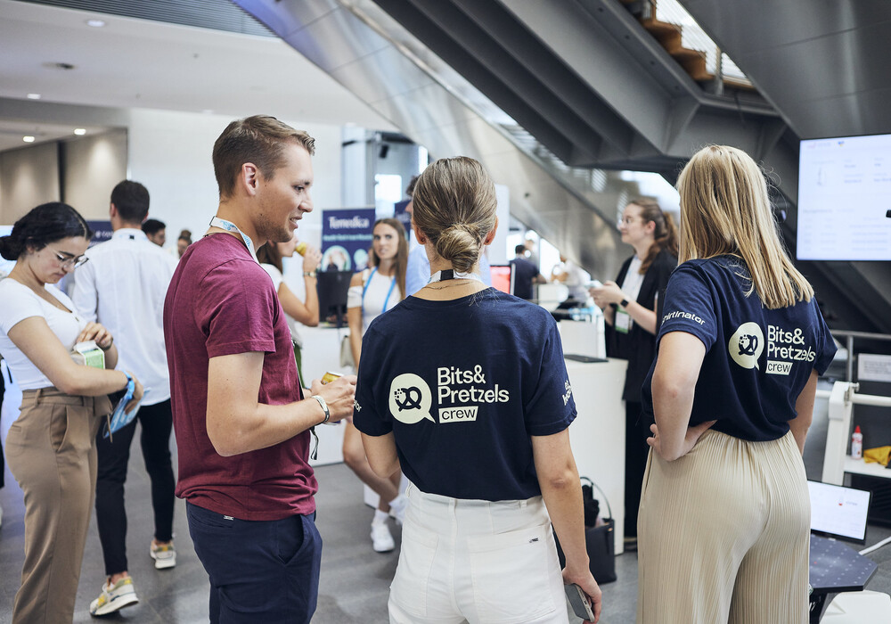 Das Bild zeigt eine lebhafte Szene auf einer Konferenz oder Messe. Im Vordergrund stehen drei Personen, zwei Frauen und ein Mann, die sich in ein Gespräch vertieft haben. Alle drei tragen T-Shirts mit dem Aufdruck "Bits & Pretzels crew". Der Mann trägt ein rotes Hemd, die Frauen beige Oberteile. Im Hintergrund sind weitere Menschen zu sehen, die sich in der hellen, modernen Halle aufhalten. Die Atmosphäre wirkt freundlich und gesellig.