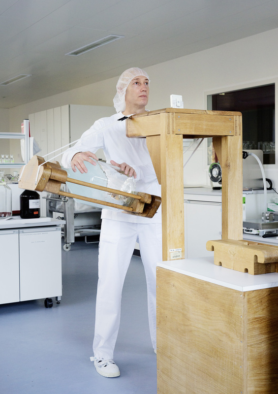 Ein Mann in einem weißen Schutzanzug und Haube steht in einem sterilen Laborraum. Er hält einen langen Holzstab mit einem Beutel darin und scheint etwas zu untersuchen oder zu bewegen. Im Hintergrund sind Laborgeräte und weiße Schränke zu sehen. Der Raum wirkt hell und sauber.