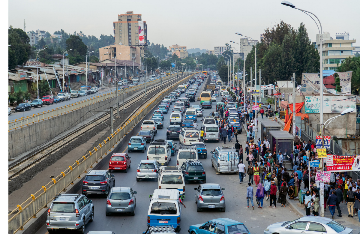 Ein breiter, städtischer Boulevard ist von dichtem Verkehr überfüllt. Autos stehen in mehreren Spuren still. Am Straßenrand versammeln sich viele Menschen, möglicherweise an einer Bushaltestelle oder einem anderen öffentlichen Ort. Gebäude säumen die Straße, und im Hintergrund sind weitere Stadtstrukturen zu erkennen. Das Ambiente wirkt geschäftig und belebt, aber auch etwas chaotisch aufgrund des Staus.