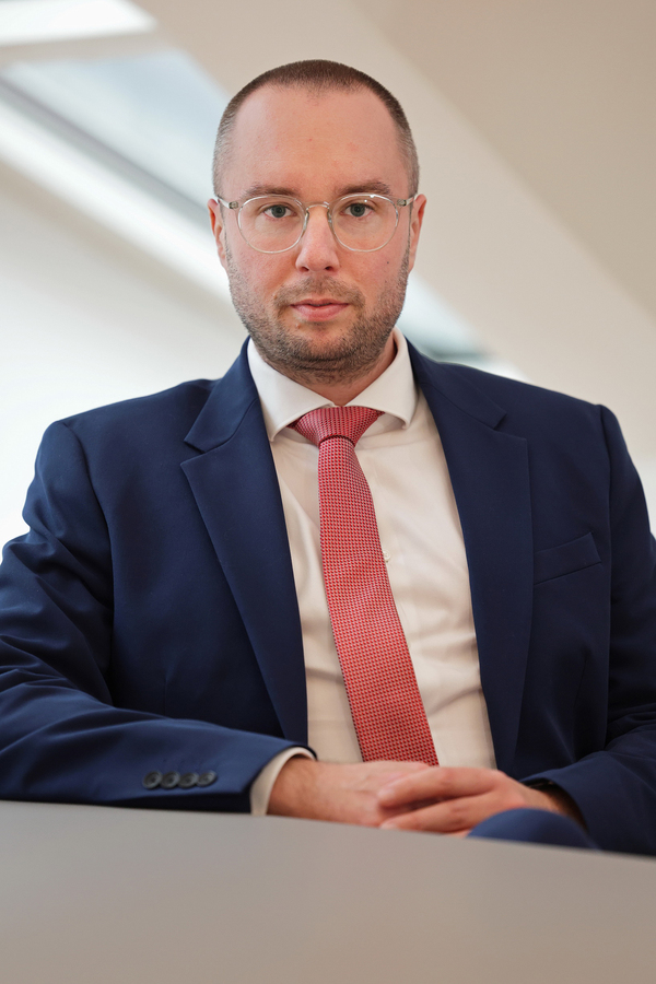 Ein Mann mit kurz geschnittenem, braunem Haar und einer Brille blickt freundlich in die Kamera. Er trägt einen dunkelblauen Anzug mit einem rot gemusterten Hemd und Krawatte. Er sitzt an einem Tisch und wirkt professionell und zugewandt.