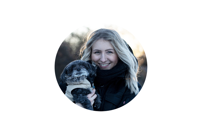 Eine junge Frau mit blonden Haaren hält einen kleinen, grauen Hund im Arm. Sie lächelt freundlich in die Kamera. Der Hintergrund ist unscharf und deutet auf einen Park oder Wald hin.