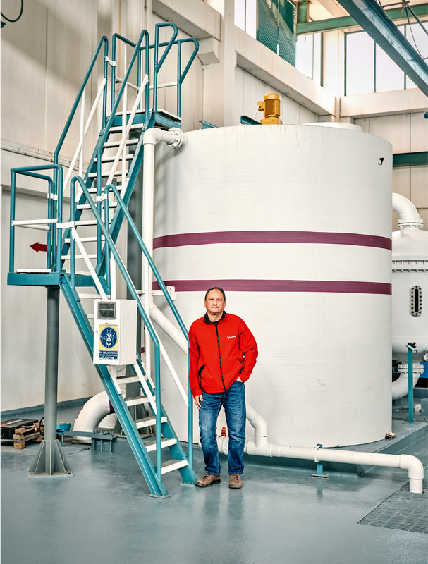 Ein Mann mittleren Alters mit kurzem Haar steht in einem großen, hellen Industriegebäude vor einem riesigen weißen Tank. Er trägt Jeans und eine rote Jacke und wirkt selbstbewusst. Im Hintergrund sind weitere Rohre und Stahlkonstruktionen zu sehen.