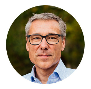 Porträt eines Mannes mittleren Alters mit grau meliertem Haar und Brille. Er trägt ein blau-weiß kariertes Hemd und blickt freundlich in die Kamera. Der Hintergrund ist unscharf und grünlich, vermutlich Vegetation.