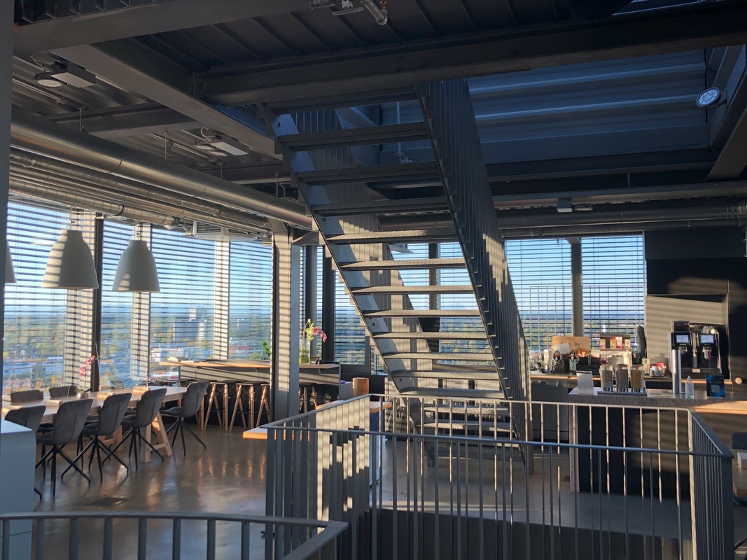 Innenansicht eines modernen Bürobereichs mit großen Fenstern und Blick auf eine Stadtlandschaft. Eine offene Treppe führt zu einer oberen Ebene. Im Vordergrund sind Tische und Stühle zu sehen, die einen Ess- oder Besprechungsbereich bilden. Das Licht scheint hell und natürlich zu sein.