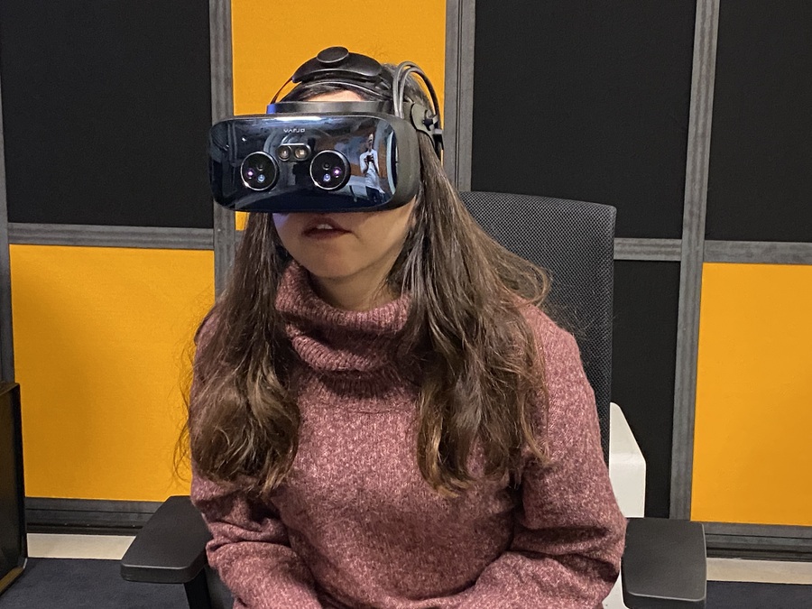 Eine Frau mit langen, dunklen Haaren trägt eine Virtual-Reality-Brille und Kopfhörer. Sie sitzt auf einem Stuhl vor einem dunklen Hintergrund. Sie scheint konzentriert zu sein.