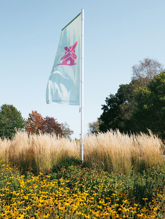 Ein großes, vertikal stehendes Banner mit einem pinkfarbenen Logo auf hellem Hintergrund ist vor einem blühenden Blumenbeet und Bäumen zu sehen. Das Banner befindet sich im Freien an einem sonnigen Tag.