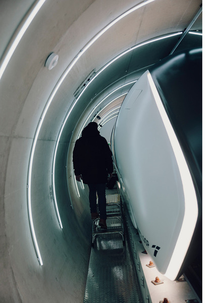Eine Person steht in einem langen, hell erleuchteten Tunnel und blickt auf ein großes, weißes, stromlinienförmiges Objekt, vermutlich ein Zug oder ein Transportmittel der Zukunft. Das Ambiente wirkt modern und technisch.
