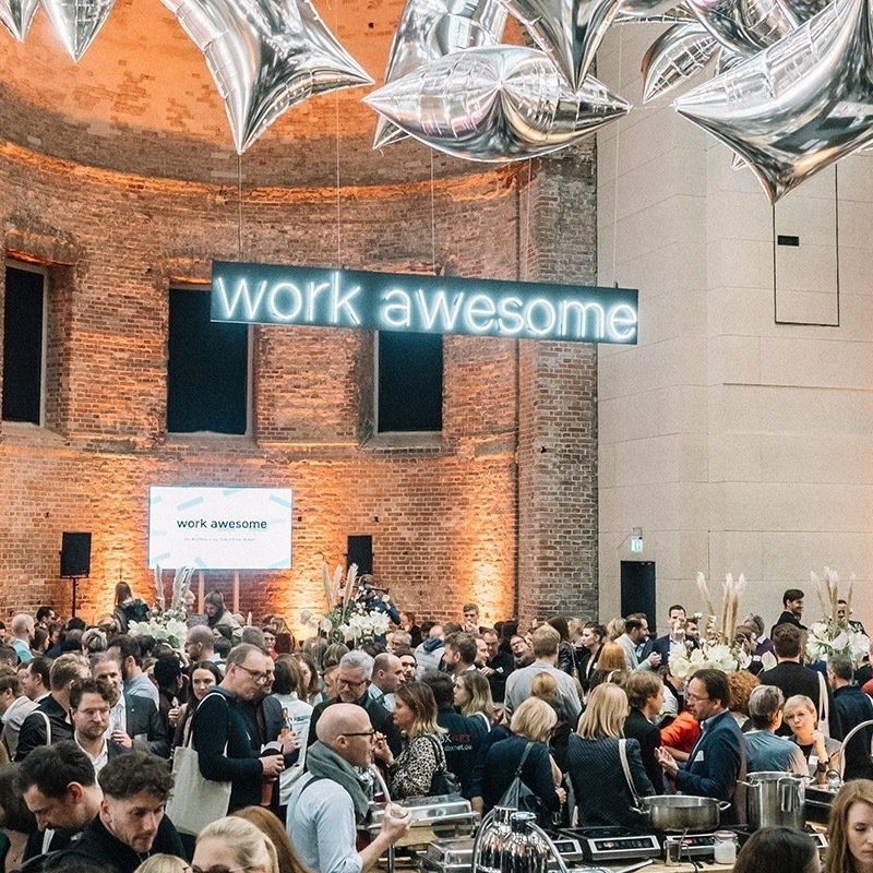 Große Menschenmenge in einem hellen Raum mit Backsteinwänden. Im Hintergrund hängt ein großes Leuchtschild mit der Aufschrift "work awesome". Menschen stehen und unterhalten sich, einige scheinen an einem Buffet zu stehen. Die Atmosphäre wirkt festlich und gesellig.