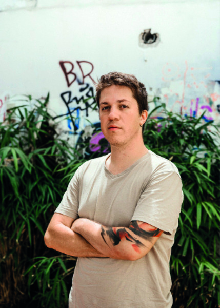 Ein junger Mann mit dunklen Haaren und einem neutralen Gesichtsausdruck steht vor einer bemalten Wand. Er trägt ein helles T-Shirt und verschränkte Arme. Im Hintergrund ist üppiges Grün zu sehen.