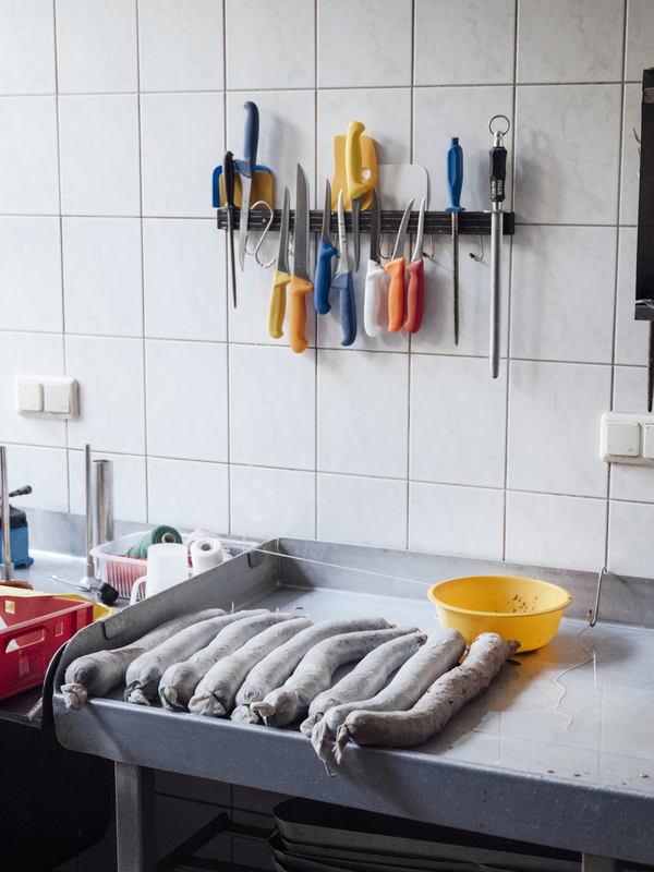 Das Bild zeigt eine Küchentheke mit verschiedenen Kochutensilien und Zutaten. An der Wand hängen mehrere Messer, auf dem Tresen steht eine gelbe Schüssel, und im Hintergrund ist ein Waschbecken zu sehen. Die Theke scheint für die Zubereitung oder Lagerung von Lebensmitteln genutzt zu werden.