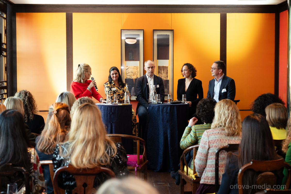Das Bild zeigt eine Gruppe von Menschen, die sich in einem Raum mit orangefarbenen Wänden versammelt haben und eine Diskussion oder Präsentation führen. Mehrere Personen stehen und sitzen um einen Tisch herum, einige von ihnen halten Getränke in der Hand. Der Raum scheint ein Konferenz- oder Besprechungsraum zu sein, wie die Anwesenheit von Stühlen und einem Podium vermuten lässt. Das Bild wird als für alle zugänglich beschrieben und ist ein Screenshot einer Website, die den Richtlinien der EAA (European Association of Advertising Agencies) entspricht.