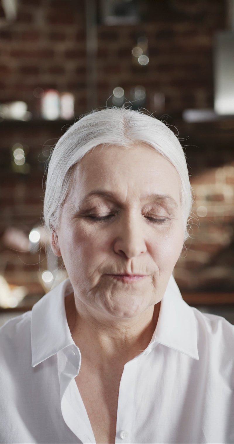 An Elderly Woman In White Shirt Breaks Into A Smile