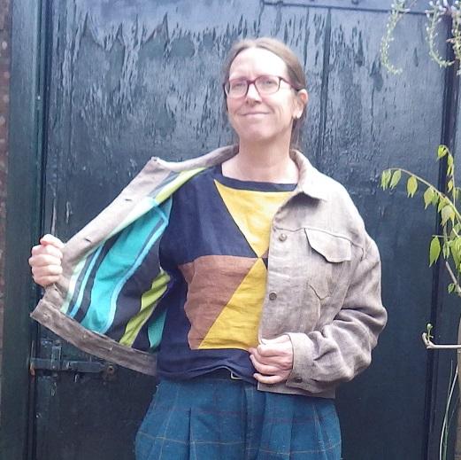 Me, wearing a short jeans style jacket in a tan linen fishbone fabric, lined with green and blue striped cotton. Under it a linen shirt with a triangle pattern in navy, brown and yellow, and some pleated green checkered trousers that look woollen, but are some dubious polyester.