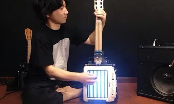 a musician sitting cross-legged holding a small old tv attached to a guitar neck; they are holding a pickup next to the screen which is displaying 5 or 6 black and white bars, and their other hand is manipulating the strings on the neck