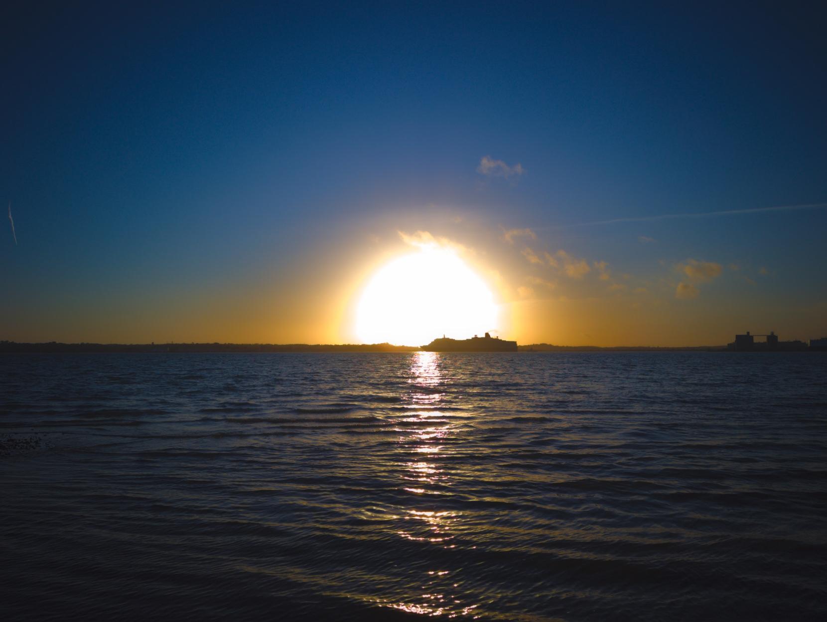 Photo of the silhouette of a cruise shipping front of the sun, right on the horizon. The sun kind of looks like a nuclear explosion.
In the foreground are the ripples of waves. In the sky there are a handful of small, fluffy clouds.
