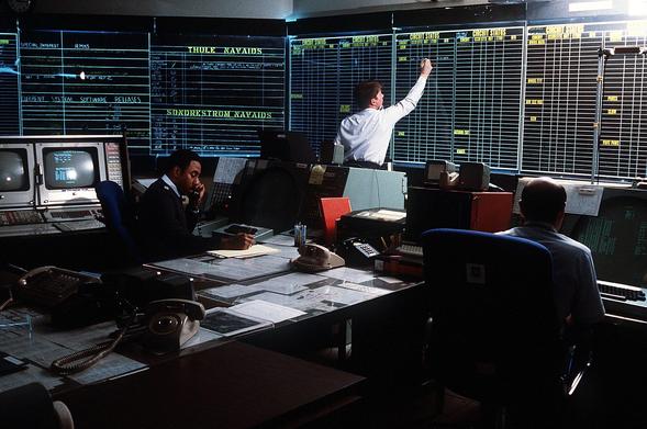 a room about the size of a school classroom with blackboards on the walls and desks covered with papers, phones, and computer terminals