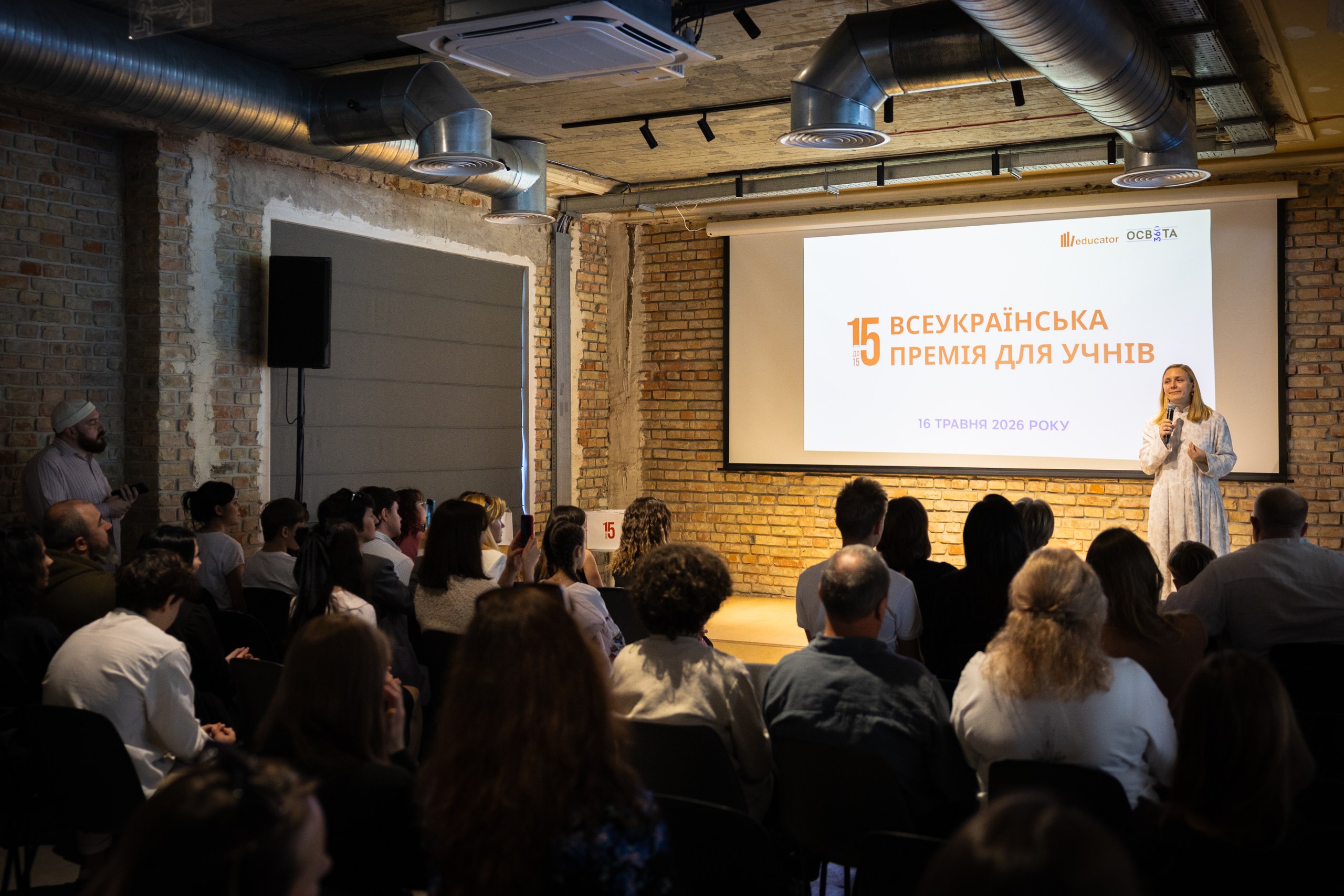 Жінка в білій сукні з мікрофоном (Владислава Бандурко, засновниця Ліцею Educator та співзасновниця ГО "Освіта 360") виступає на сцені на тлі цегляних стін поруч із проєкційним екраном із заголовком "15 Всеукраїнська премія для учнів", 16 травня 2026 року, та логотипами "educator" і "Освіта.ua". Ліворуч на сцені — чоловік із мікрофоном у руці. У переповненому залі сидять і стоять глядачі, дехто фотографує подію на телефон