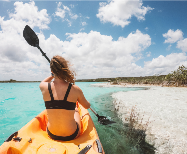 Vrouw in kajak met de rug naar de camera toe in bikini en met peddel vast op het water met uitzicht op de vlakte
