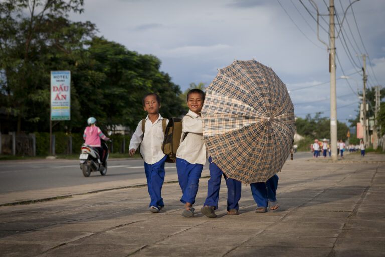 4 Vietnamese kinderen lopen over straat, ze dragen een grote paraplu met zich mee