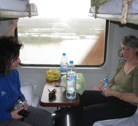 twee vrouwen zitten tegenover elkaar in de trein, ze drinken uit grote waterflessen en boven hen zie je een klein stukje van twee bedden