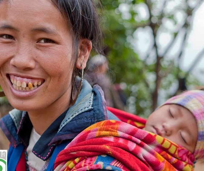 Vietnamese vrouw kijkt lachend de camera in, ze heeft een slapend kindje in een draagzak op haar rug