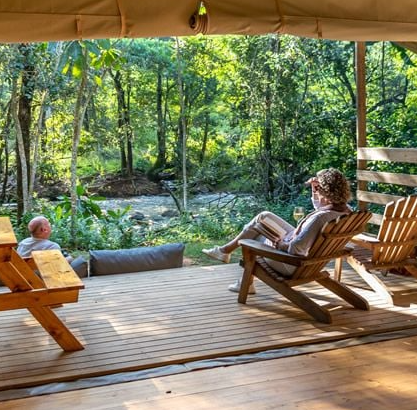 Reiziger met glas wijn in stoel op de veranda van een glamping tent en een andere reiziger zittend voor de veranda met uitzicht op de rivier en de bomen