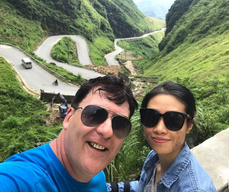 koppel kijkt lachend in de camera met op de achtergrond de kronkelende wegen van de Ha Giang Loop