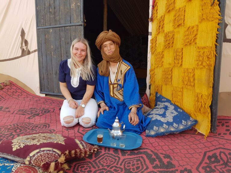 een toerist zit naast een vrouw in traditionele kleding op een kleed, ze hebben een dienblad met thee voor zich