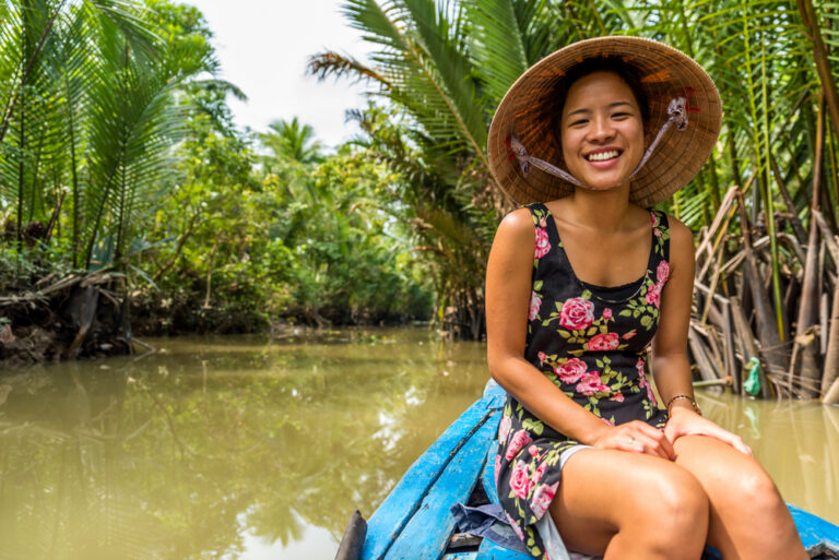 een vrouw op de punt van een bootje, ze kijkt lachend de camera in en heeft een Vietnamese hoed op haar hoofd