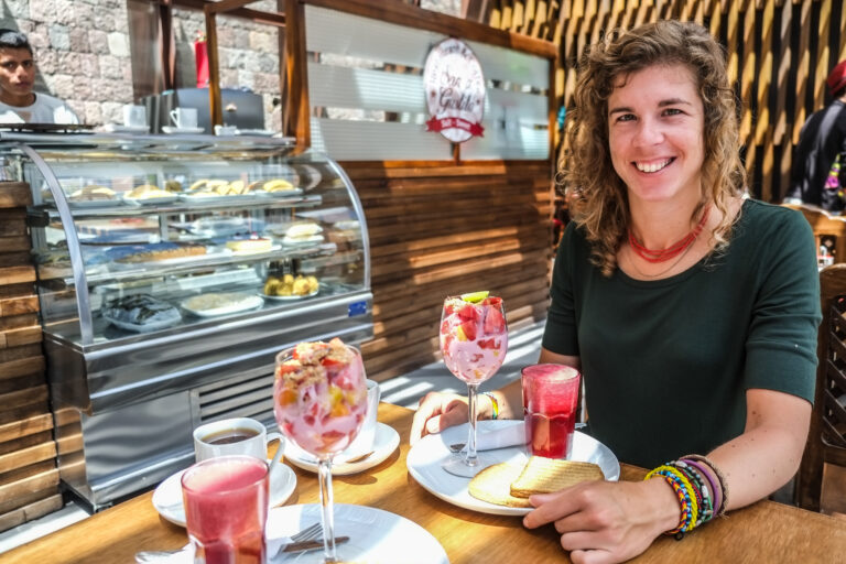 vrouw zit in een café met een ontbijtje voor zich: koffie, toast, yoghurtbowl en sap