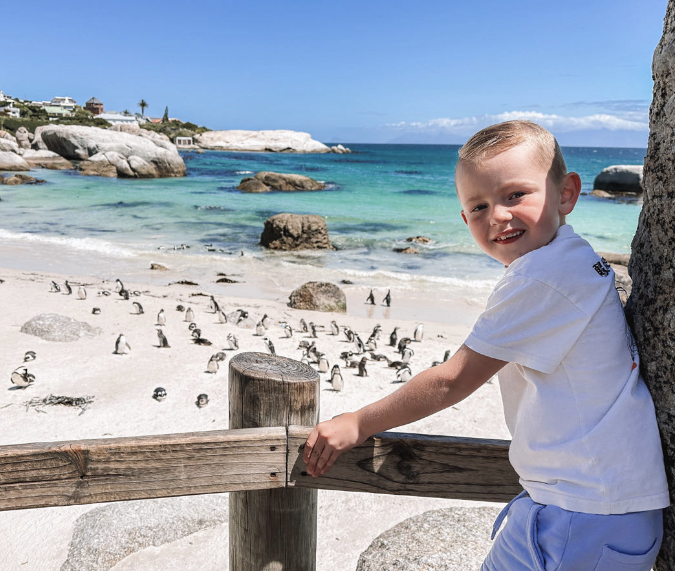 Jongen lachend in de camera met op de achtergrond een strand met pinguins en de zee in Zuid-Afrika