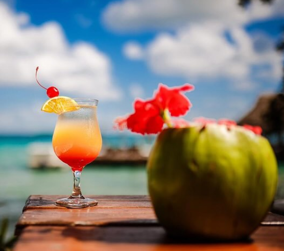 Een cocktail met sinaasappelschijf en kers op het glas en vaag op de voorgrond een kokosnoot met bloem