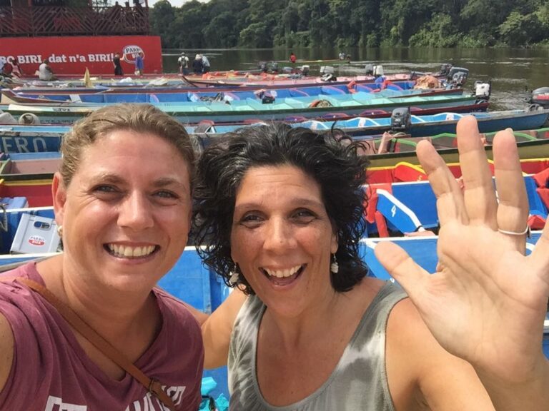 twee vrouwen zwaaien met hand met op achtergrond korjaal boten op Surinamerivier
