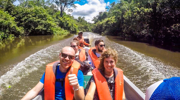 een selfie van 4 toeristen in een bootje op een rivier waar langs groene bomen staan