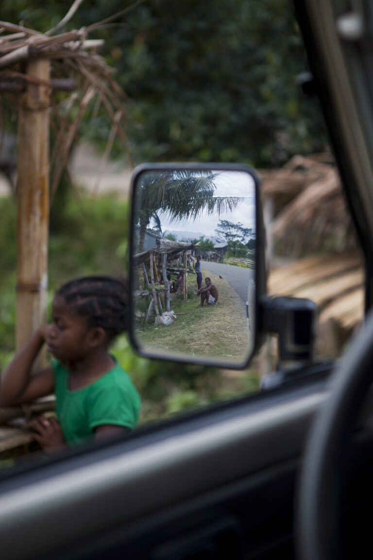 Weg en local vanuit auto gefotografeerd in autospiegel met op de achtergrond een lokaal kindje in Madagaskar