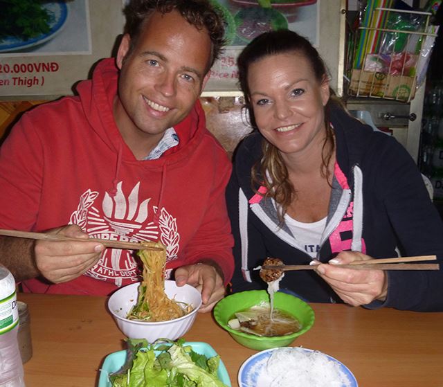 koppel kijkt lachend de camera in, in hun handen allebei stokjes waarmee ze noodles uit hun kommetje eten
