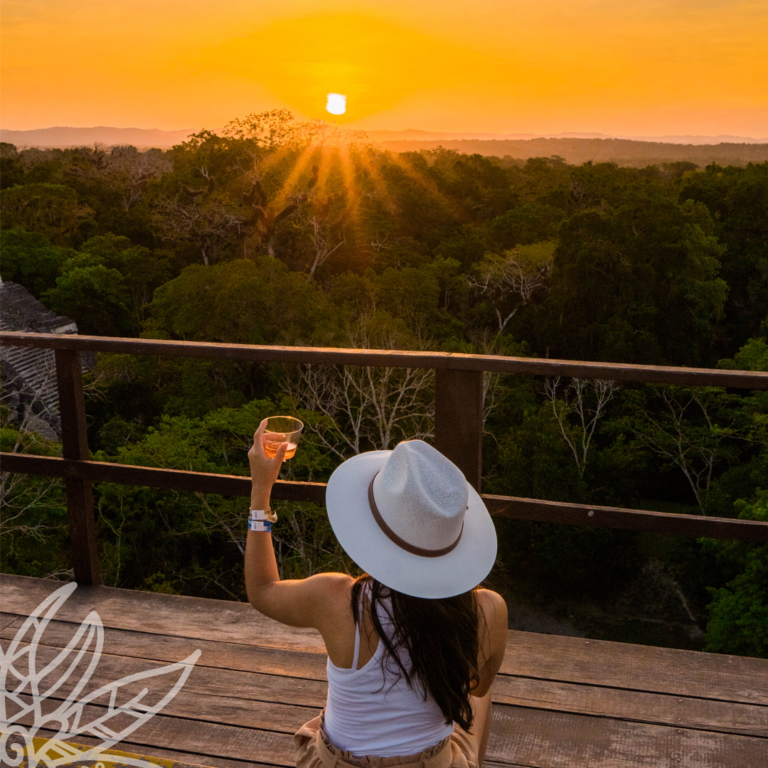 Vrouw met hoed en met de rug naar de camera proostend met een drankje en uitkijkend op de jungle en zonsondergang