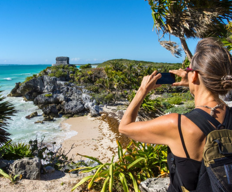 Vrouw met de rug naar de camera en een foto makend met haar mobiel van een Maya ruïne aan het strand in Mexico in Tulum