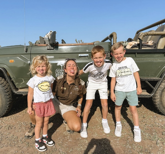 3 kinderen staan op de foto met een ranger, ze steken alle vier hun tong uit naar de camera en staan voor een jeep