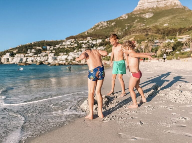Drie kinderen staand in badkleding op het strand bij de zee met op de achtergrond een centrum en heuvel