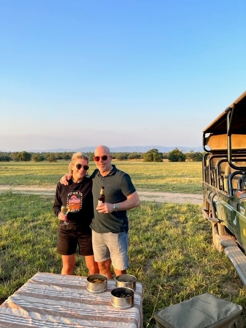een koppel staat naast een truck op de savanne met een drankje in de hand voor een zonsondergang tour