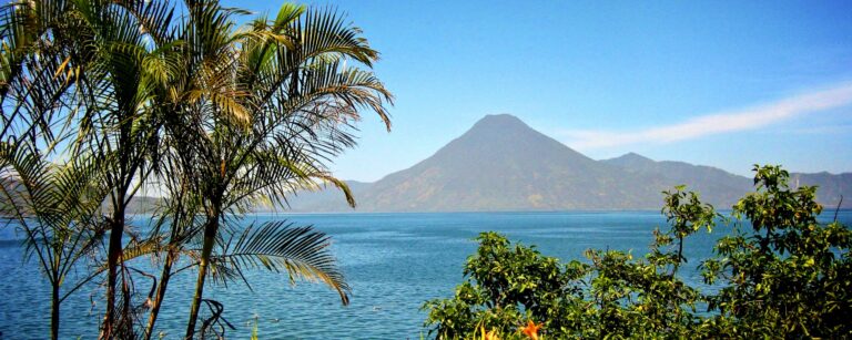 Groot meer met op de achtergrond een vulkaan en op de voorgrond palmbomen en meer groen in Guatemala