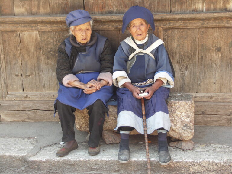 twee oude aziatische vrouwen in Naxi klederdracht zitten op een stenen bankje voor houten gebouw in China