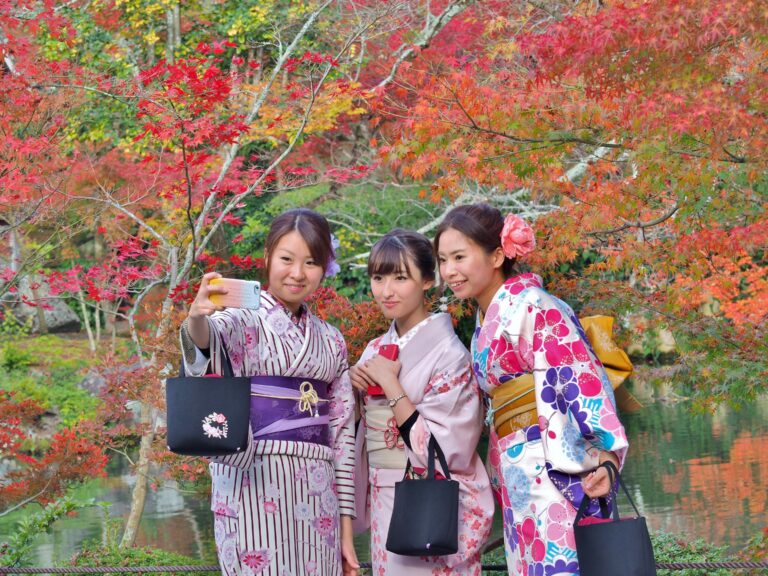 drie dames gekleed als geisha in kleurrijke kimono's maken selfie omringd door kleurrijke bloesembomen
